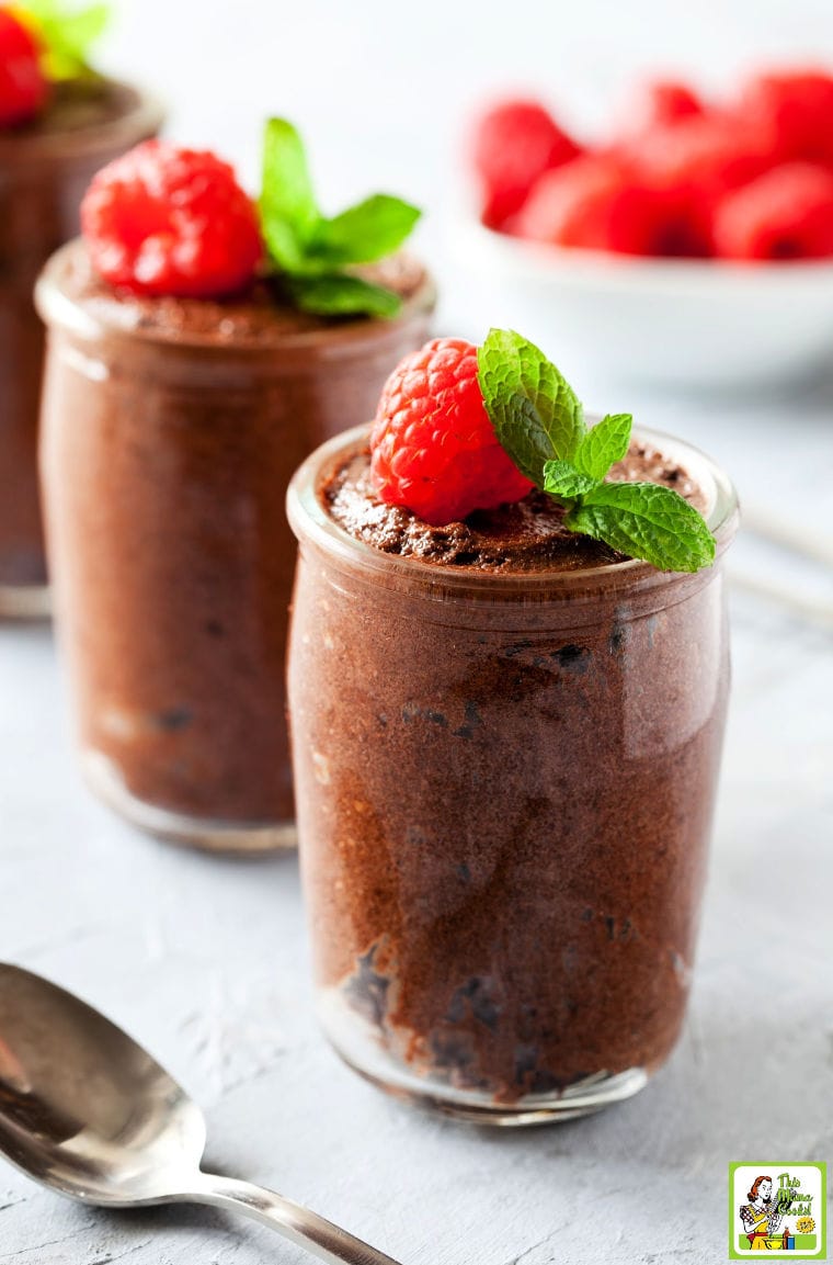 Two dessert glasses of chocolate mousse served with garnish of raspberries and mint. With a bowl of raspberries in the background and a spoon.