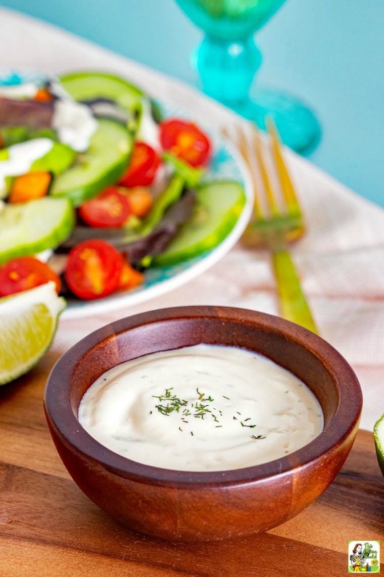 A cup of Dill & Lime Vegan Dressing Recipe with a plate of salad, tomatoes and lime on a wooden cutting board.