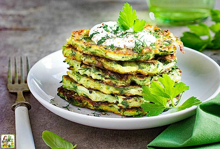 A white plate with Zucchini Fritters topped with a dollop of sour cream and some fresh herbs.