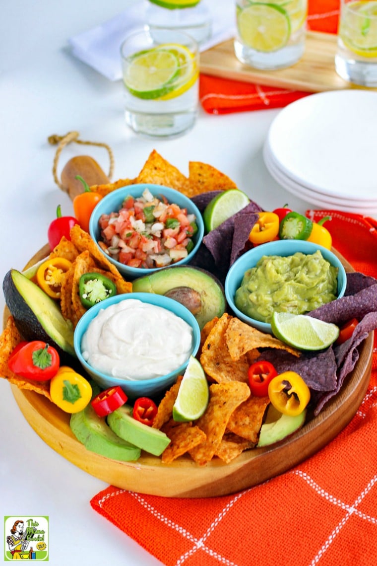 Overhead view of a platter with a small blue bowl of sour cream with slices of limes, tortilla chips, mini sweet peppers, avocados, blue bowls of salsa and guacamole, with white plates and glasses of water with slices of limes and lemon.