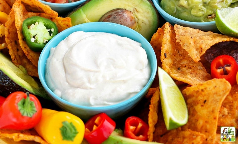 A small blue bowl of vegan tofu sour cream with tortilla chips, mini peppers, avocado, guacamole, and bowls of salsa.