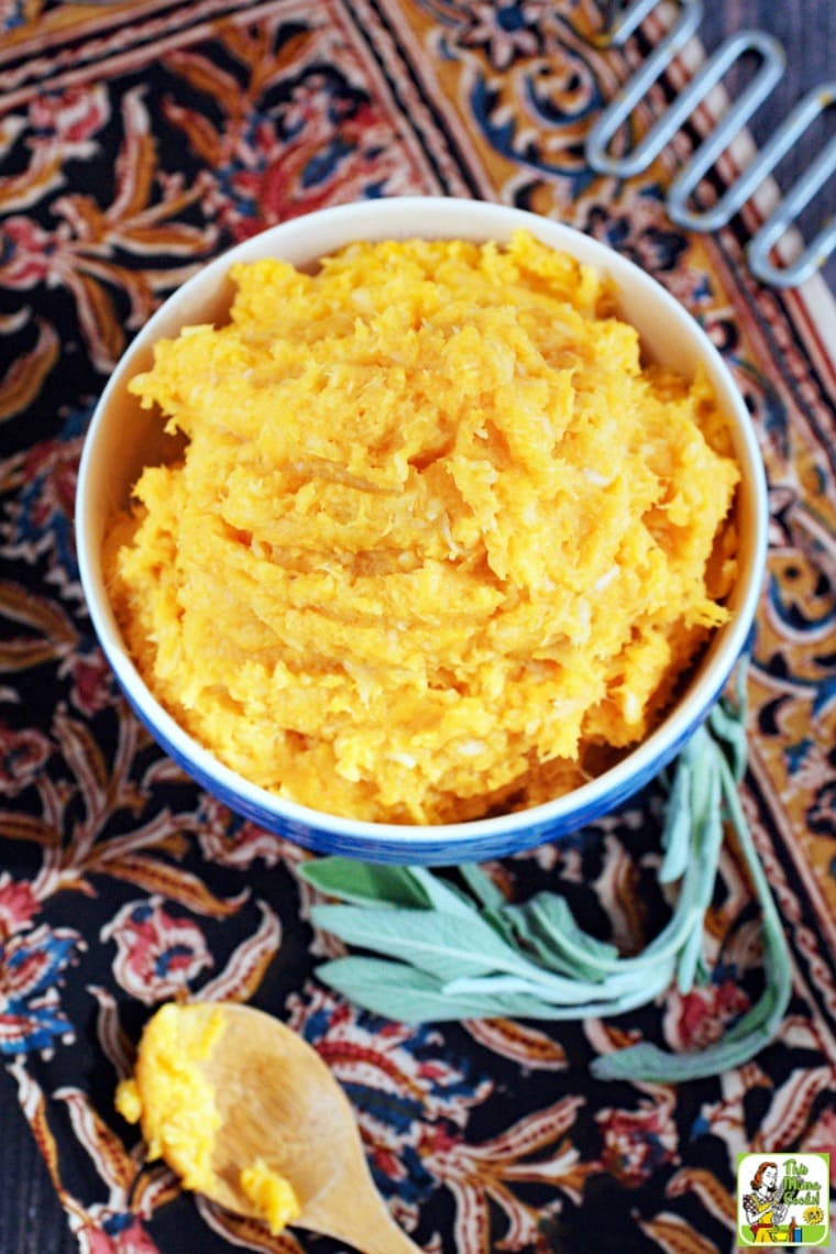 A blue bowl of Mashed Sweet Potatoes with an antique potato masher, a sprig of sage, and wooden spoon.