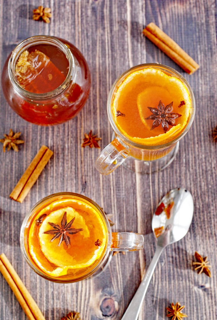 Overhead view of mugs of hot toddy with tea, spices, cinnamon sticks, spoon, and a glass jar of honey.