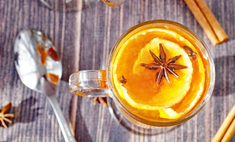 A glass mug of hot toddy with tea with orange peel, spoon, cinnamon stick, and spices.