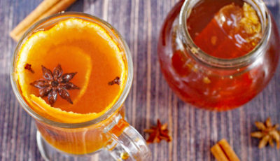 Glass mug of hot toddy with orange peels, star anise and cloves with a jar of honey and spices.