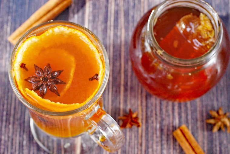 Glass mug of hot toddy with orange peels, star anise and cloves with a jar of honey and spices.