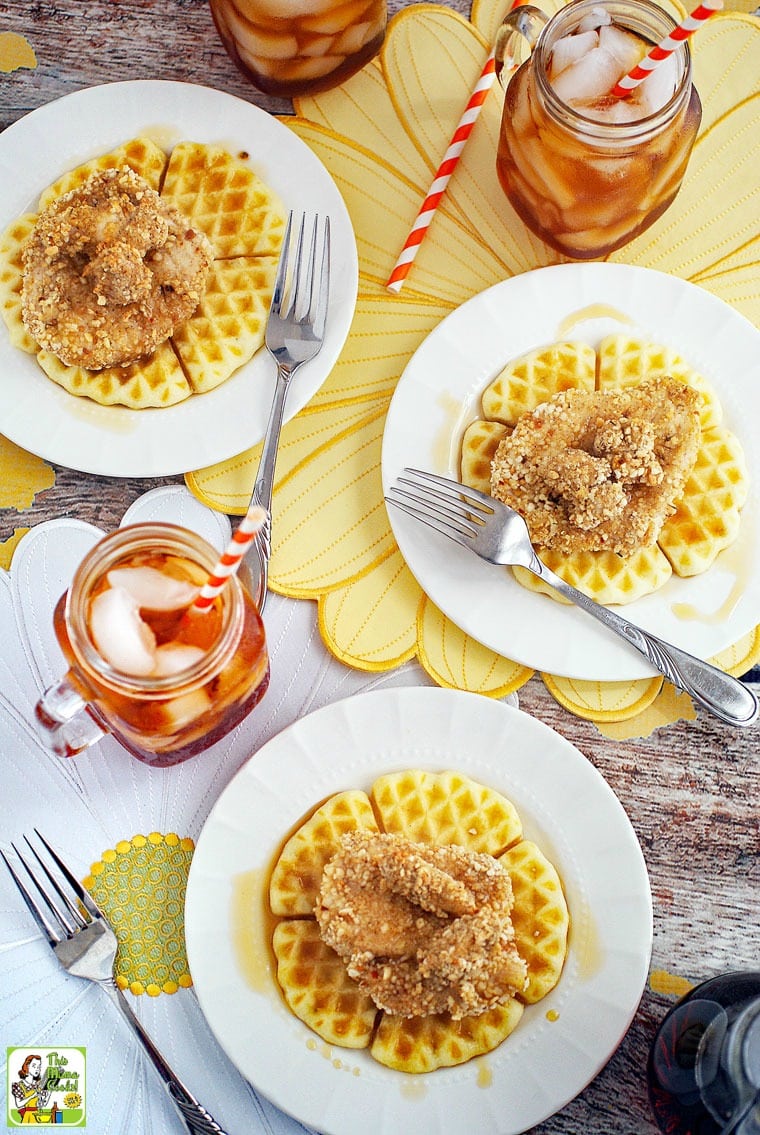 Overhead view of baked chicken and waffles covered on a white plates with forks, glasses of ice tea, and straws, on white and yellow floral placemats.