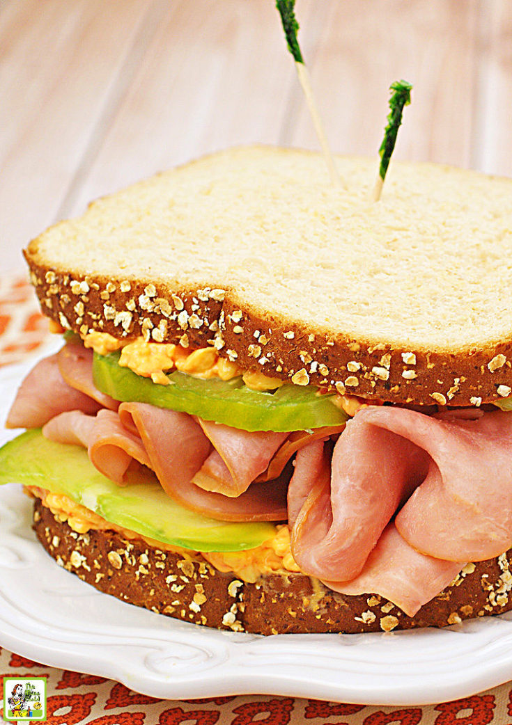 Ham & Pimento Cheese Sandwich with a green heirloom tomato on a white plate with an orange and white napkin.