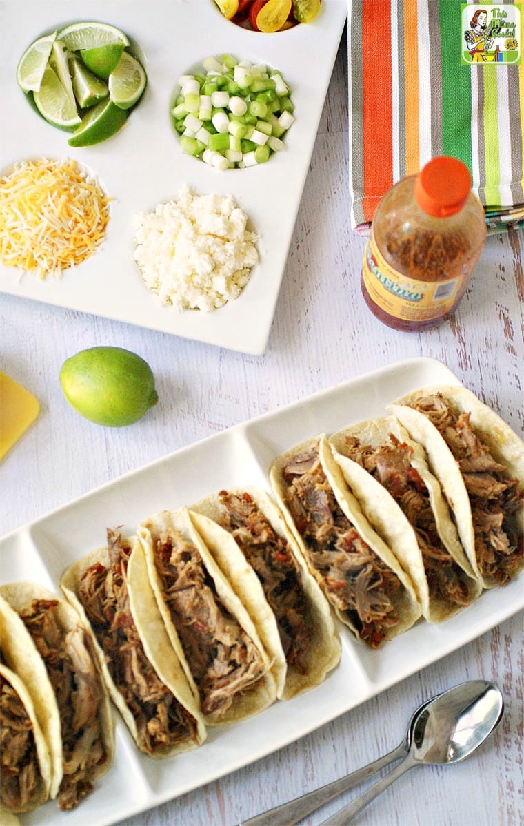 A platter of pulled pork tacos with a tray of toppings, a lime, and hot sauce.