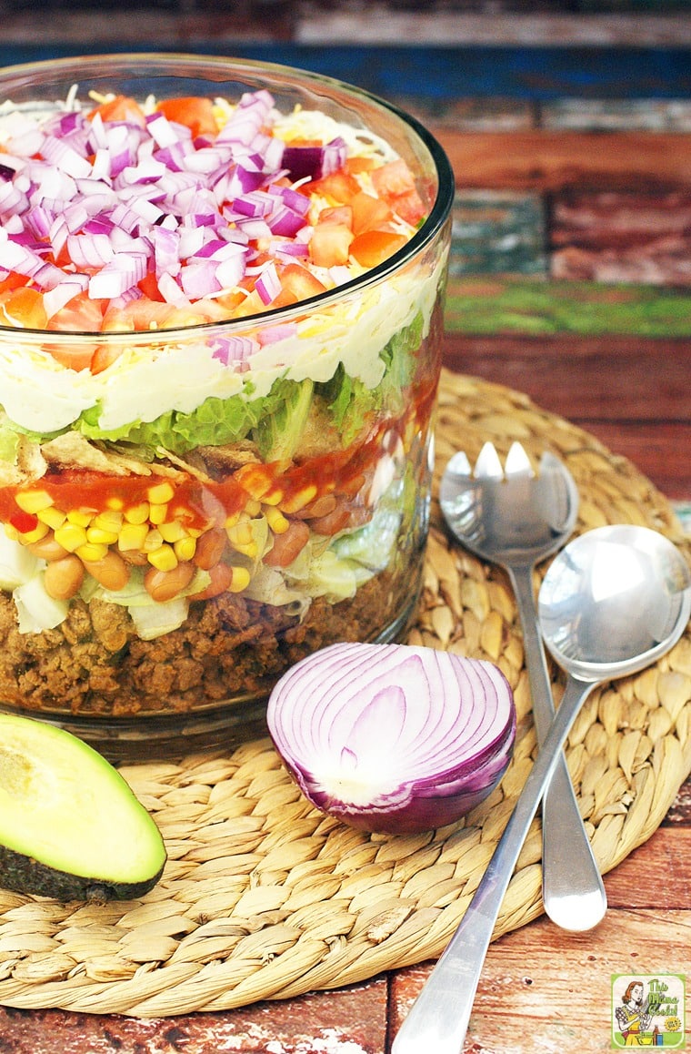 Taco salad in a trifle bowl with onion, avocado and serving utensils.