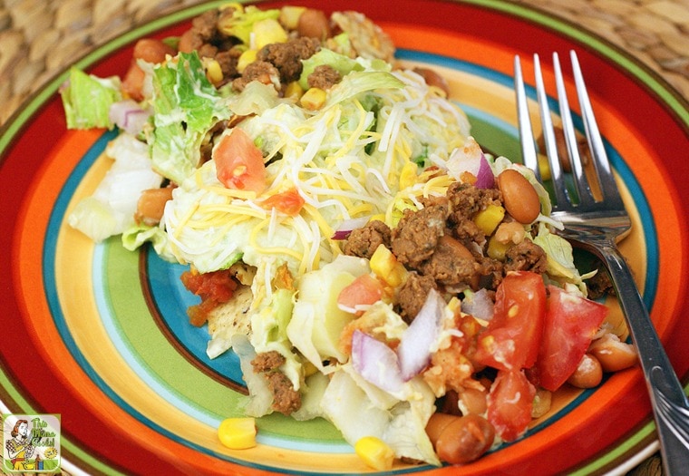 A colorful striped plate of taco salad with beans, tomatoes, salad, corn, lettuce, and cheese served with a fork.