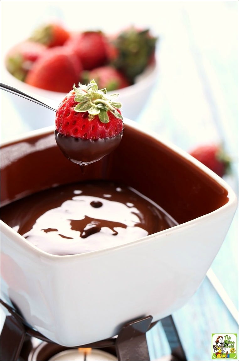 Eine Erdbeere auf einem Fondue-Spieß taucht in eine Tasse Schokoladenfondue mit einer Schüssel Erdbeeren im Hintergrund.