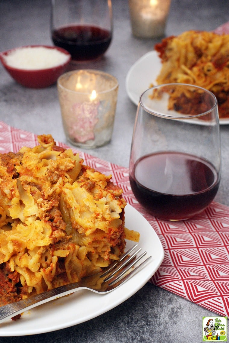 A plate of slow cooker baked ziti on a white plate with silver fork with a red napkin with a glass of red wine with a candle, another plate of ziti, and a small bowl of shredded cheese.