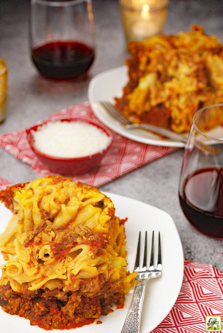 Plates of slow cooker baked ziti on white plates with forks, red napkins, a small red bowl of shredded cheese, and glasses of red wine.