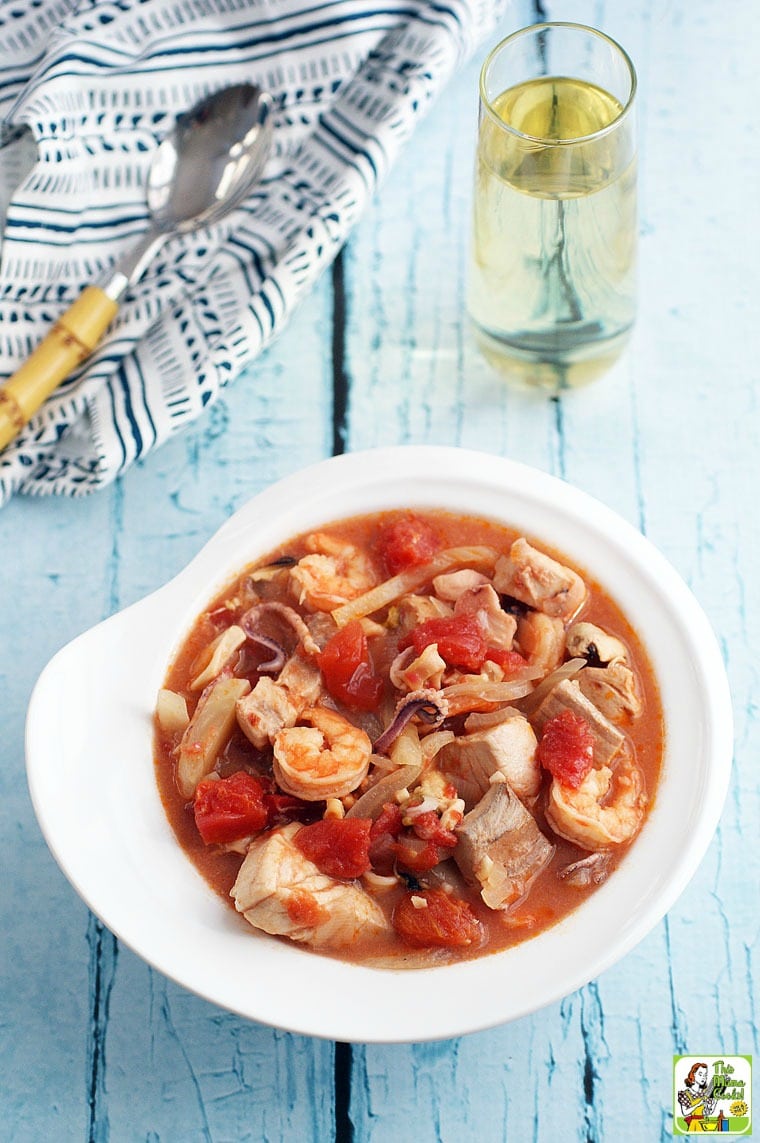 A bowl of tomato-based cioppino seafood stew. Wine glass, spoon and napkin in the background.