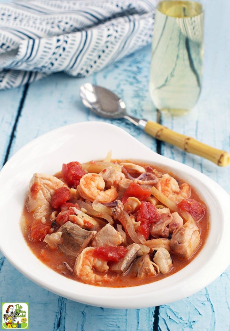 Closeup of a bowl of tomato-based Italian seafood stew. Wine glass, spoon and napkin in the background.