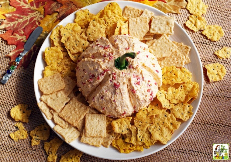 A large white plate with crackers and pimento cheese spread pumpkin on an autumn leaf placemat with a jeweled knife.