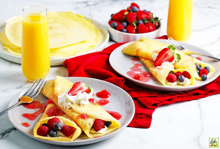 Plates of rice flour crepes with raspberries, blueberries, whipped cream, and berry drizzle with red napkins and glasses of orange juice.