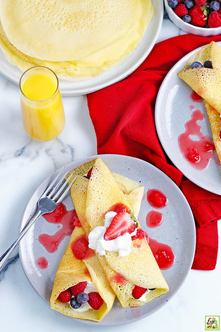 Overhead shot of two gluten free crepes stuffed with berries and covered in whipped cream and strawberry drizzle with napkins, and other plates of crepes, and a glass of orange juice.