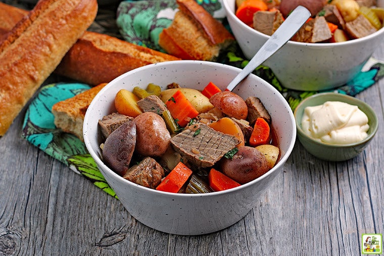 Slow Cooker Venison Stew in a white bowl with a spoon next to a small bowl of horseradish and rolls of crunchy bread.