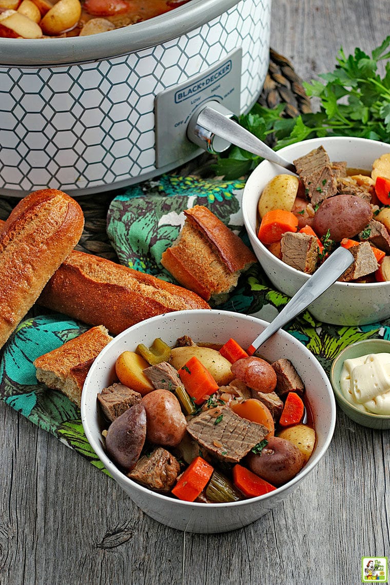White ceramic bowls of venison and vegetable stew with spoons, colorful napkins, bread rolls, a small bowl of horseradish, and a slow cooker in the background.