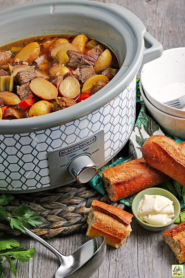 A slow cooker of venison stew with vegetables and potatoes with a ladle, chunks of bread, a bowl of horseradish, and plates in the foreground.