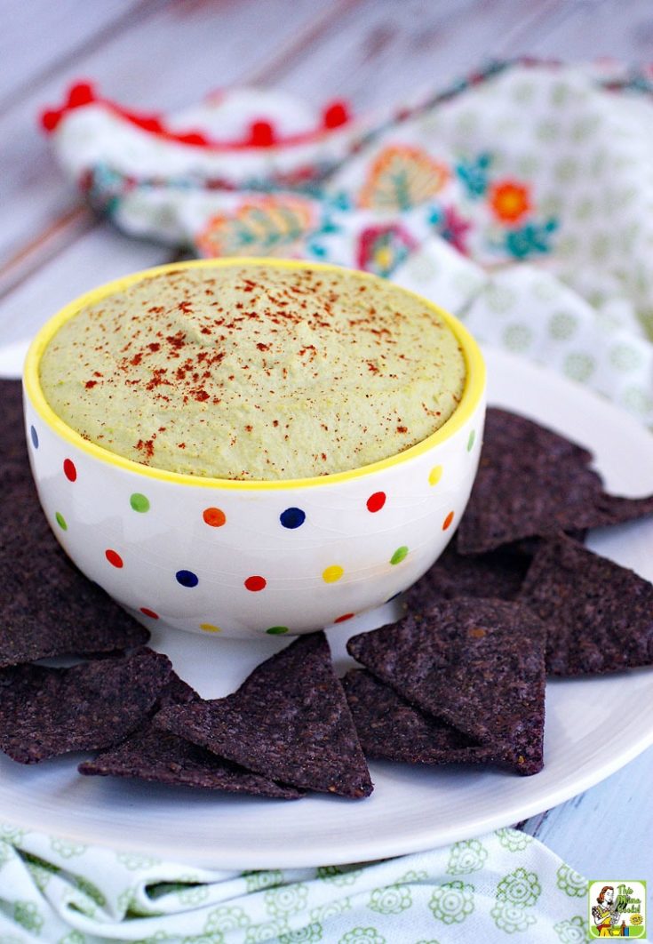 A white bowl with polka dots with green edamame hummus on a white dish with blue tortilla chips.