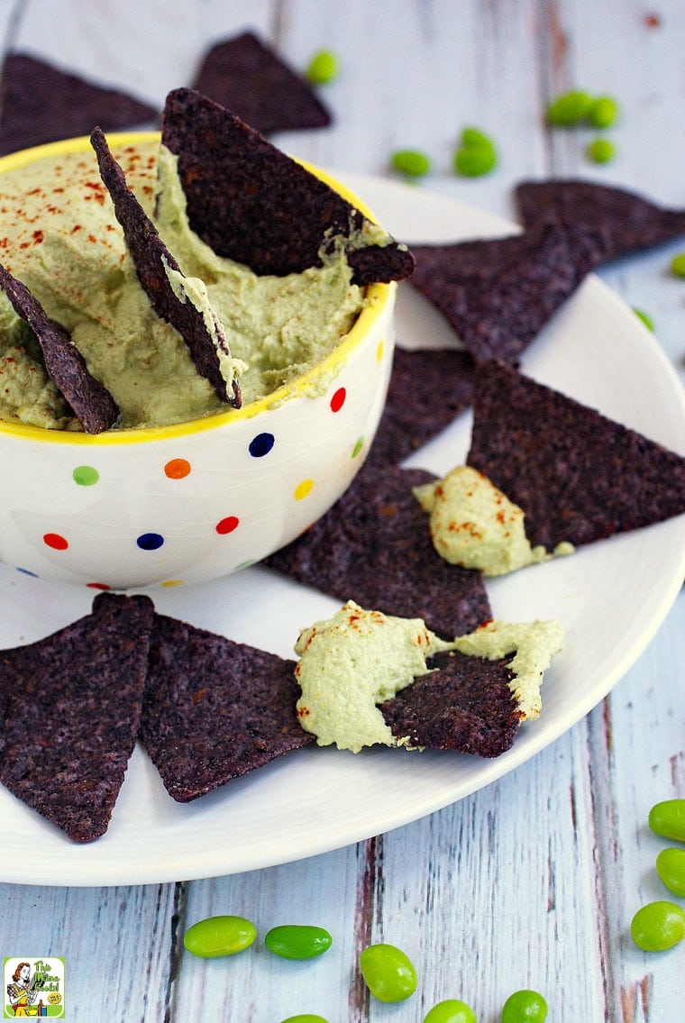 A white bowl with polka dots with blue tortilla chips dipped into green edamame hummus placed on a white plate.