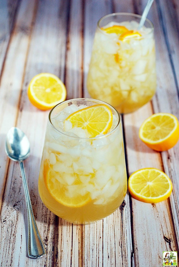 Two cocktail glasses of lemon shrub drinks on ice with sliced lemons and spoon.