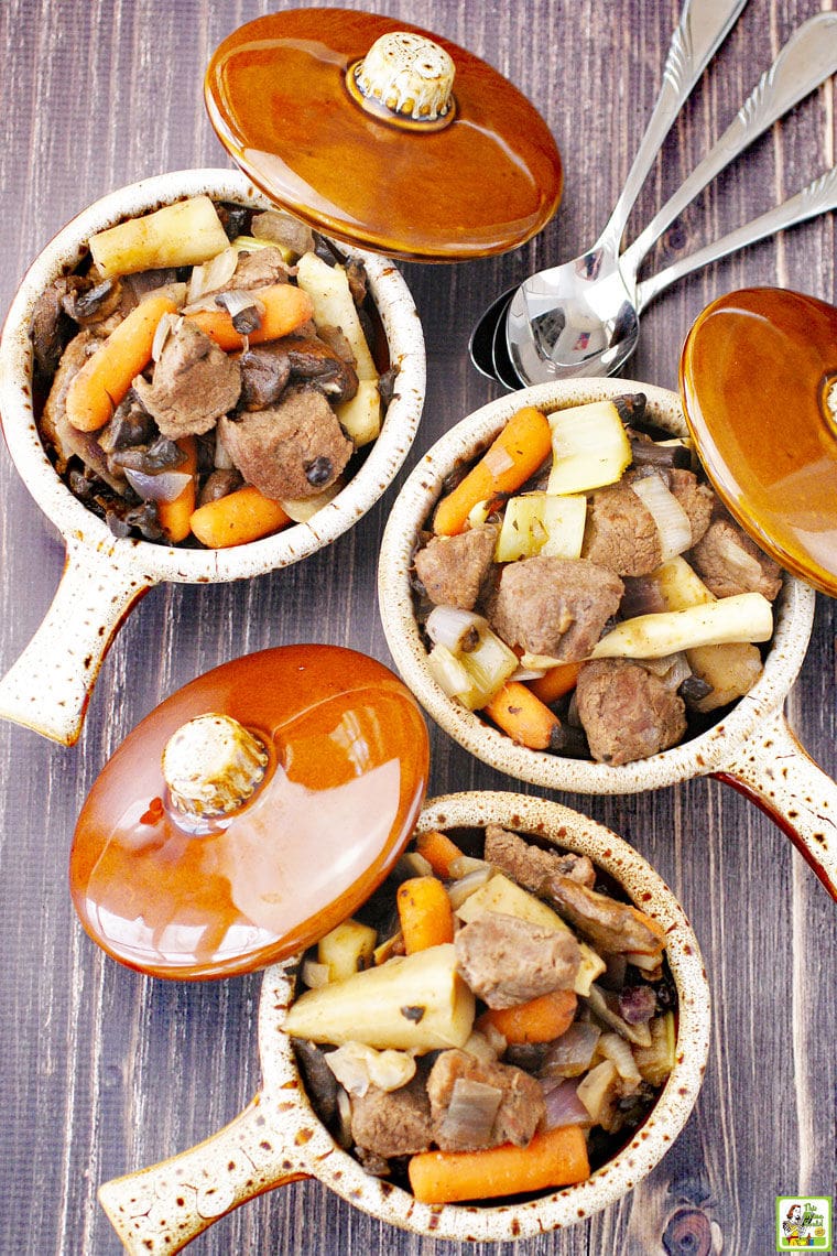 Overhead view of three brown bowls with lids of crockpot deer stew filled with carrots, parsnips, onions, and mushrooms and silver spoons.