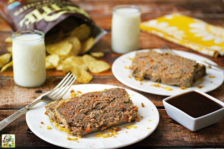 Two white plates of meatloaf with a glasses of milk, forks and napkins, a bag of potato chips, and extra BBQ sauce.