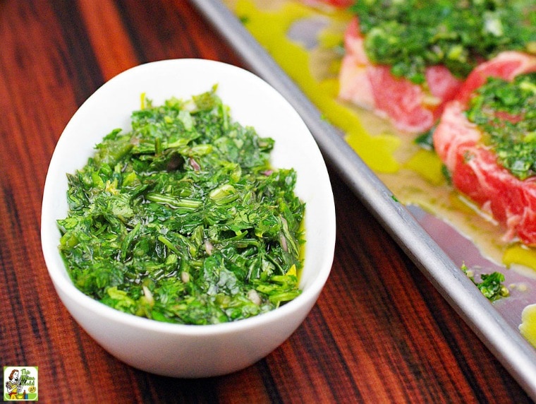 A bowl of Chimichurri Marinade and Sauce with a tray of steaks covered in chimichurri.