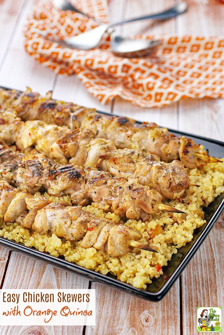 Chicken Skewers served on quinoa a black platter with serving utensils and an orange napkin in the background.