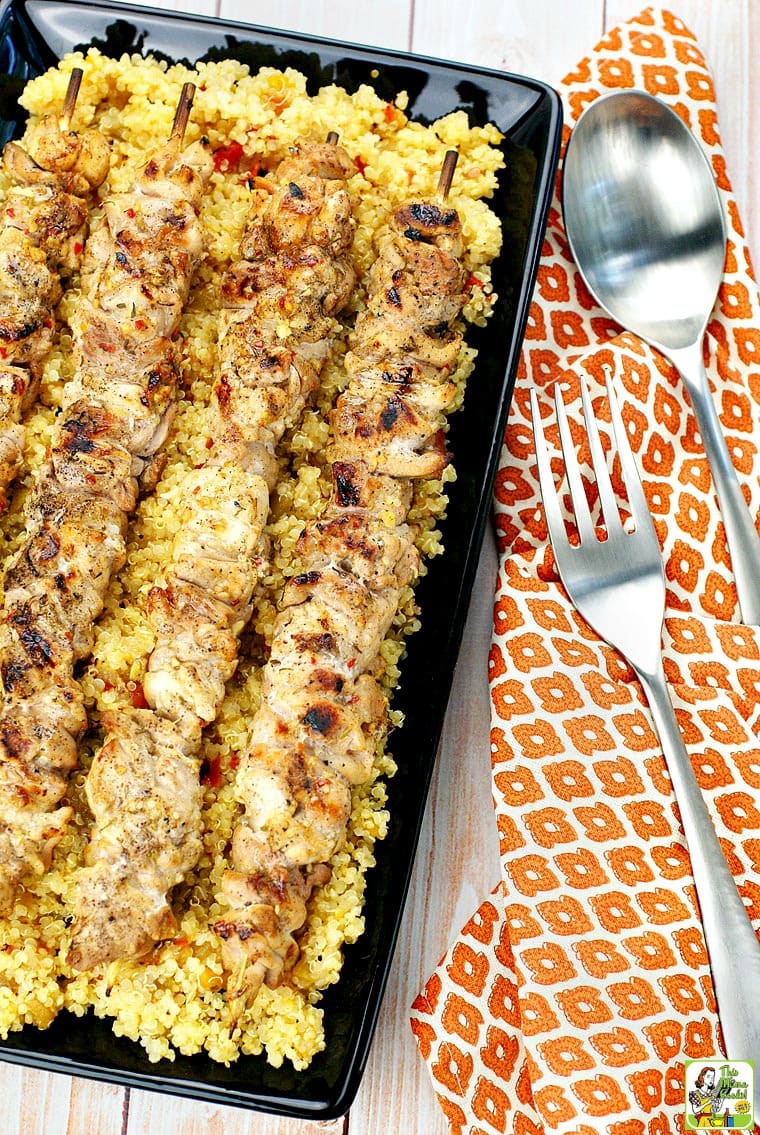 Chicken Skewers served on Quinoa a black platter with serving utensils and an orange napkin.