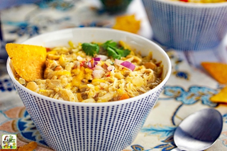 A blue bowl with slow cooker white chicken chili served with tortilla chips, chopped onions, and parsley with a spoon.