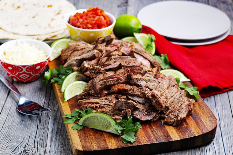 Sliced carne asada meat with limes and cilantro on a wooden cutting board with taco fixings.