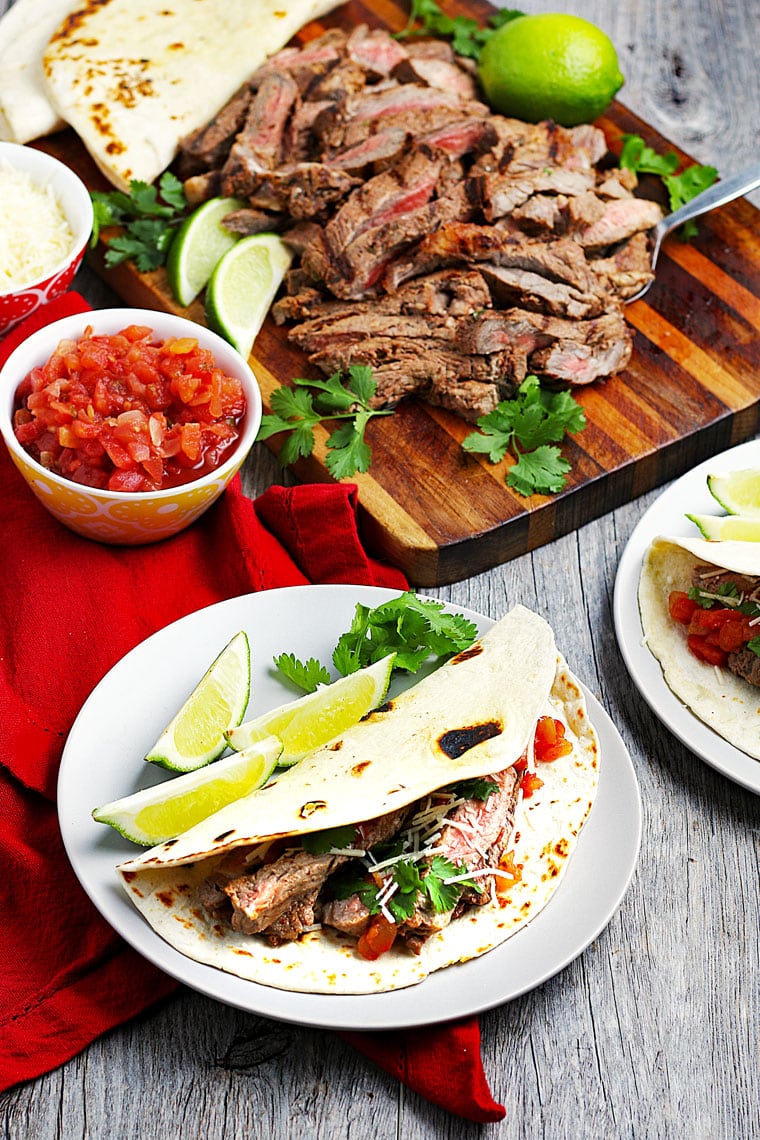 Grilled steak tacos on plates, sliced meat with limes and cilantro on a wooden cutting board, and a bowl of salsa.