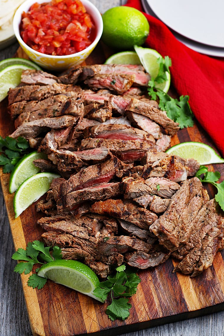 Sliced grilled meat with limes and cilantro on a wooden cutting board and a bowl of salsa.