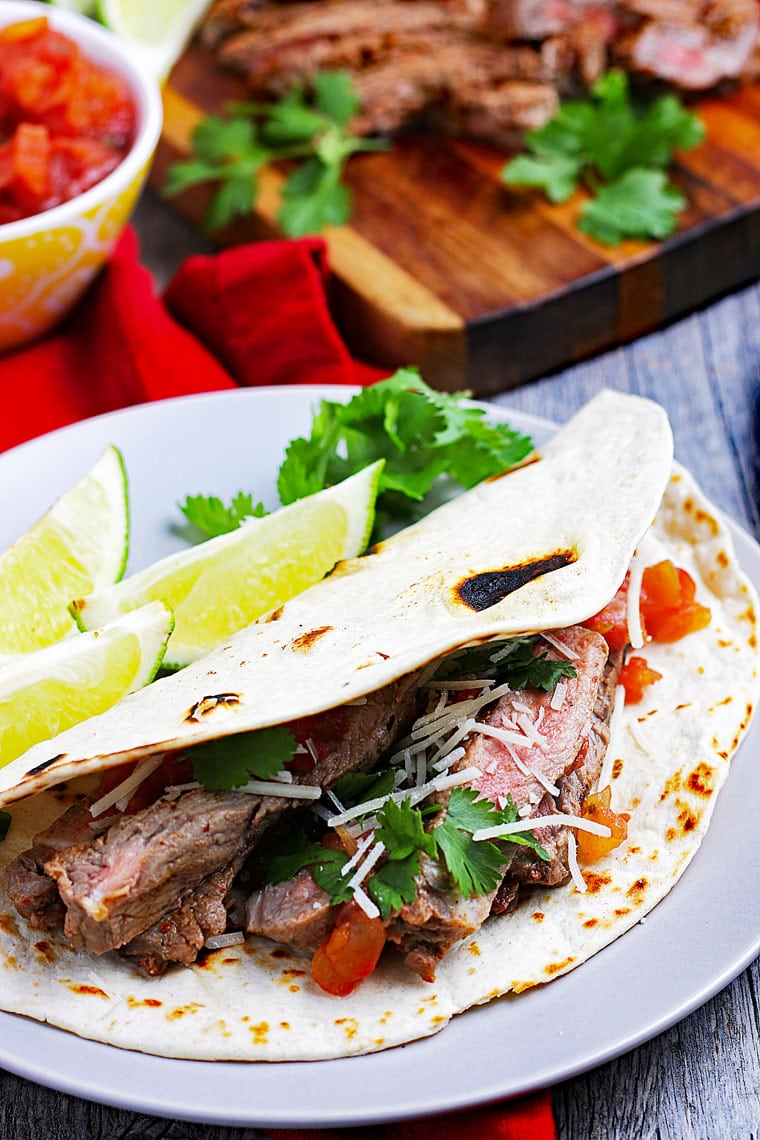 Closeup of a carne asada taco on a white plate with limes with a bowl of salsa and grilled meat on a cutting board.