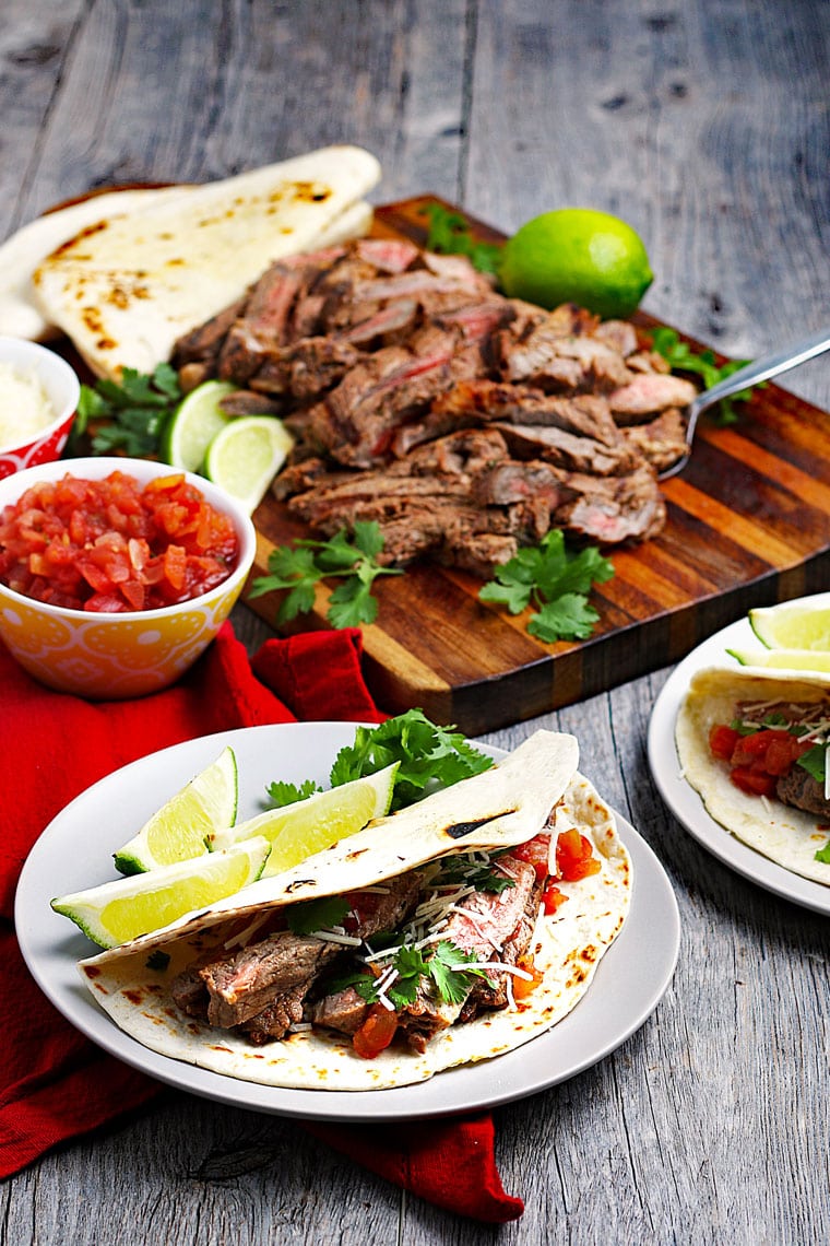 Plates of tacos with sliced meat on a cutting board with a bowl of salsa and plates of tortillas.