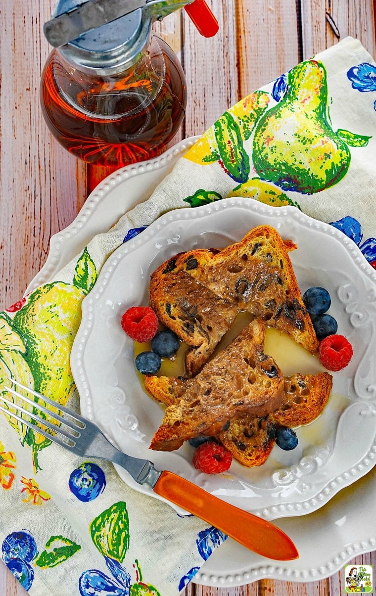 A white plate of French Toast with berries and fork on a colorful napkin with a pitcher of maple syrup