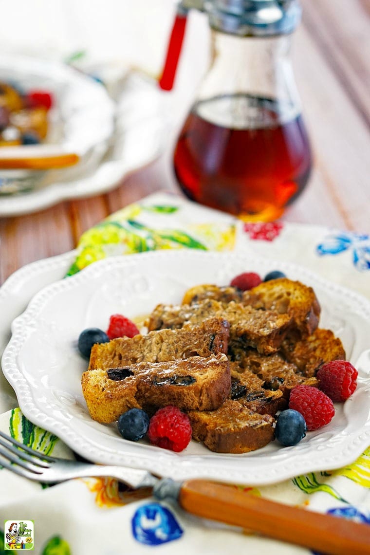A white plate of French Toast with berries with a pitcher of maple syrup in the background