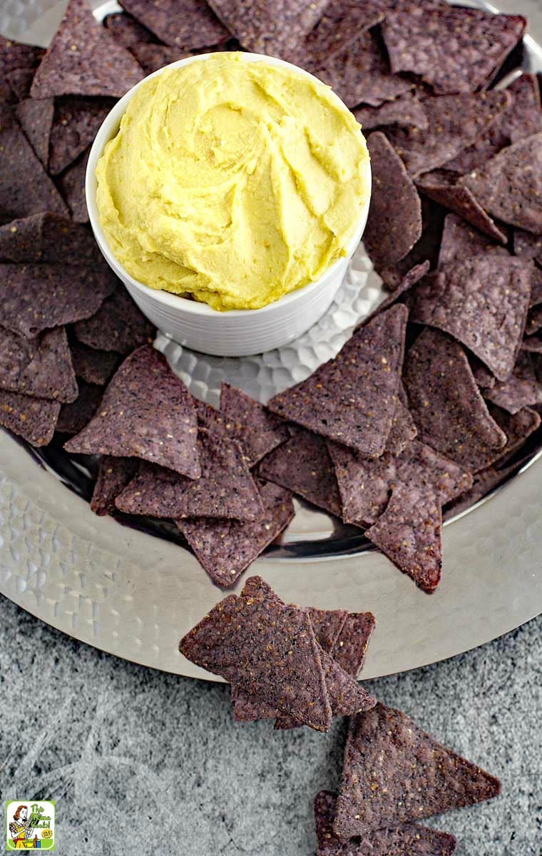 Overhead shot of avocado hummus dip in a white bowl with  blue tortillas chips on a silver tray.