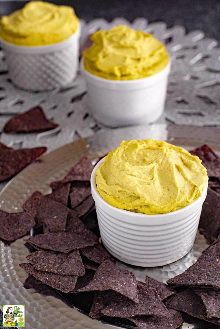 White bowls of avocado hummus with  blue tortillas chips on a silver tray.