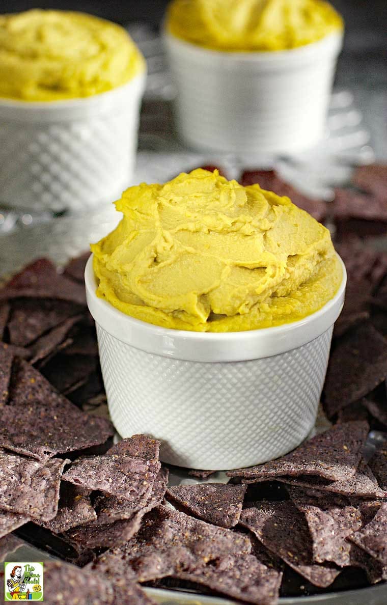 A white bowl of avocado hummus with  blue tortillas chips on a silver tray.