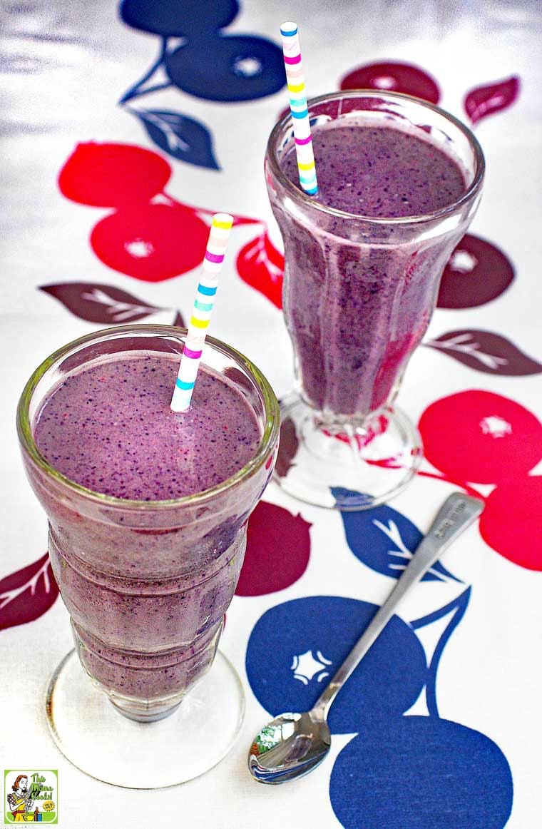 Two milk shake glasses filled with berry smoothies served with colorful striped straw and a serving spoon on a fruit patterned tablecloth.