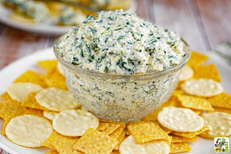 A glass bowl of Vegan Spinach Artichoke Dip with crackers on a white plate.