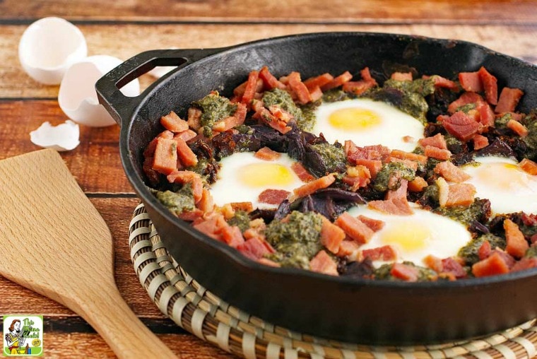 Baked eggs, ham, potatoes, sun dried tomatoes, and pesto in a skillet with a wooden serving spoon.