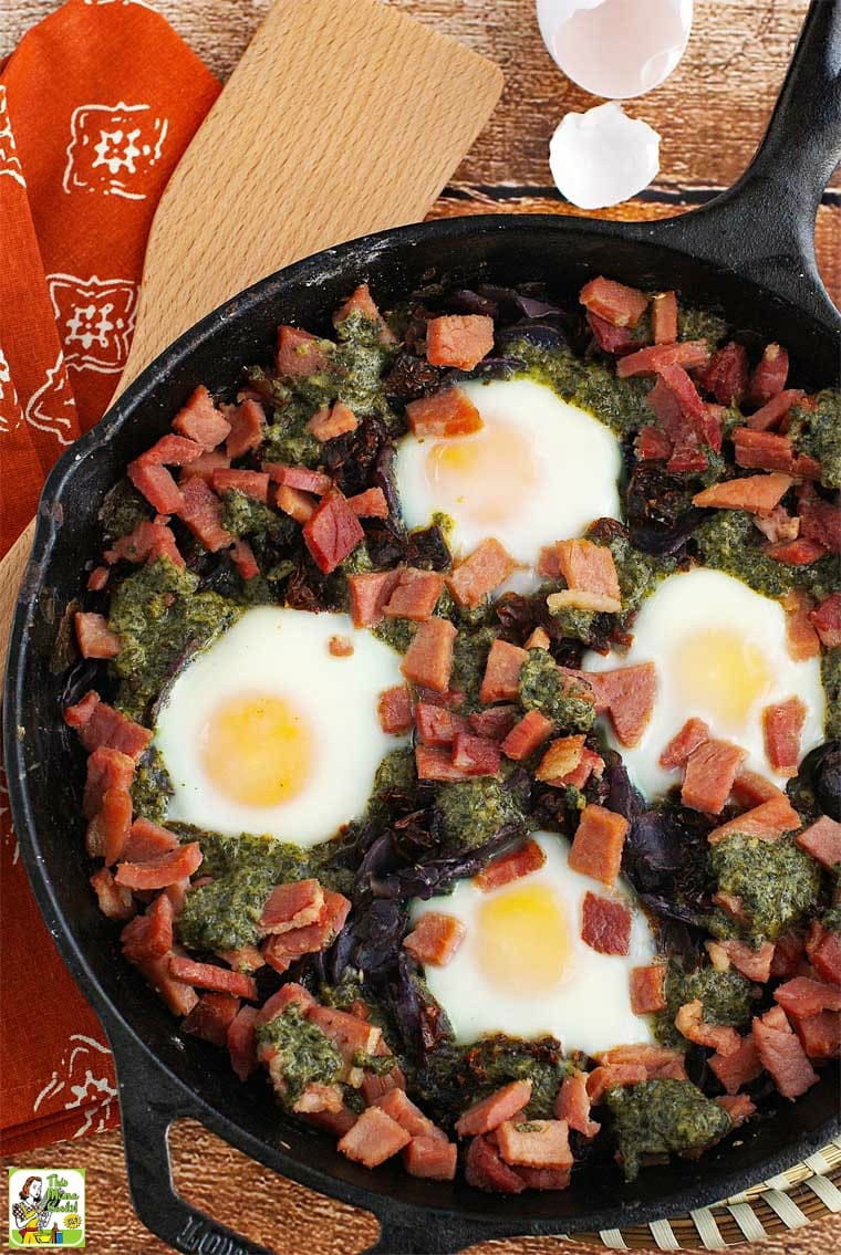 Overhead shot of baked eggs, ham, sun dried tomatoes, potatoes, and pesto in a cast iron skillet.