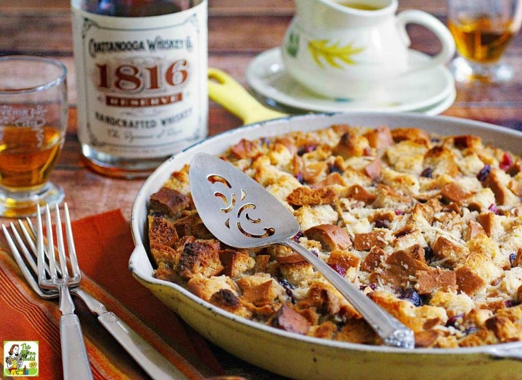 A skillet of Bread Pudding with Whiskey Sauce with serving knife, forks, bourbon bottle and glass, and pitcher of sauce.